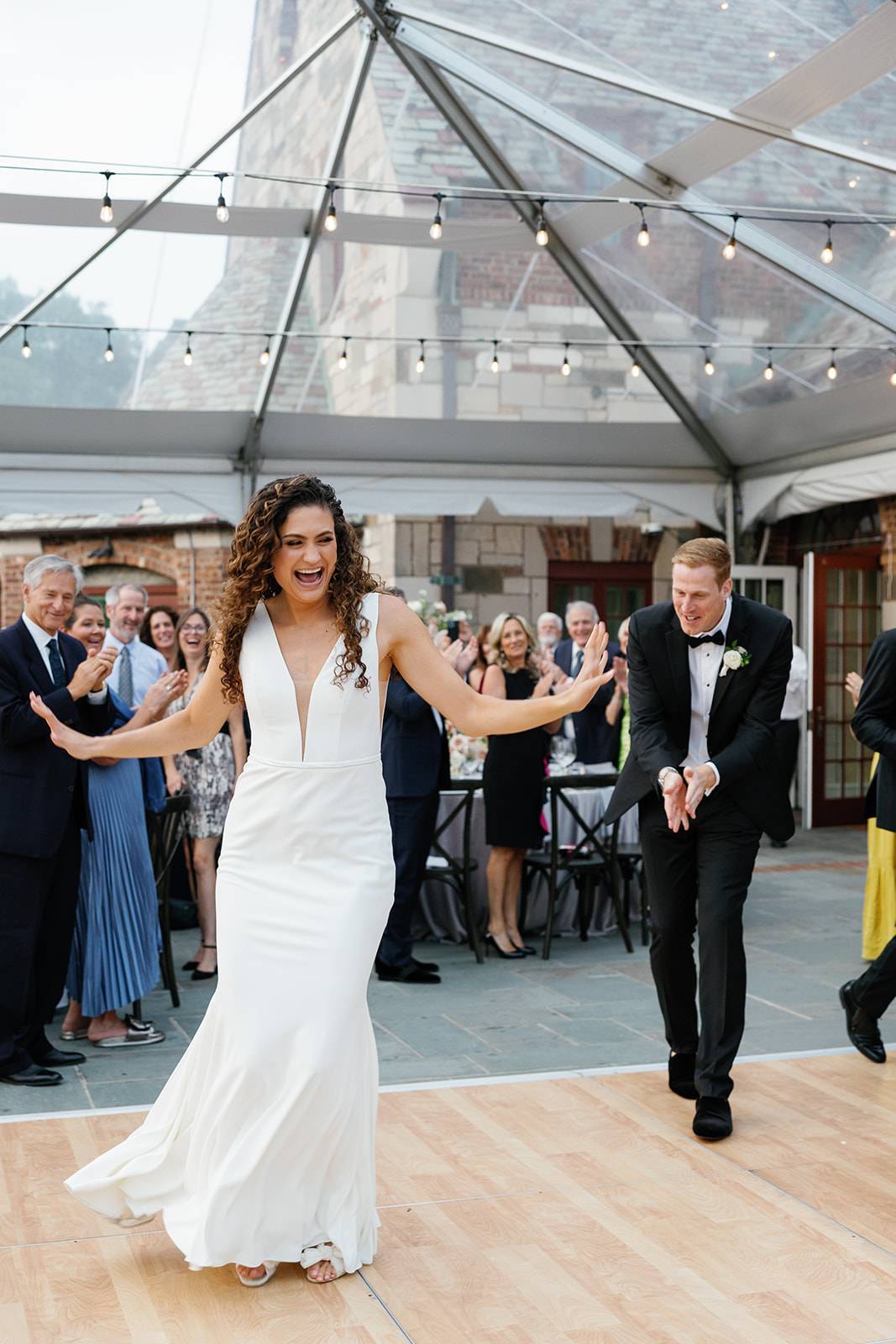 A very happy and smiling bride and groom dancing on the dance floor at their wedding reception under a clear top tent with string bulb lights.